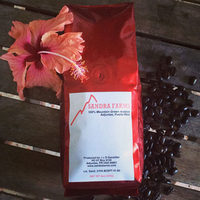 Red coffee bag from Sandra Farms with coffee beans and a pink flower on a wooden surface